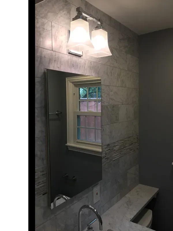 Bathroom with gray tile, a mirror, and a window. A two-light fixture is mounted above the mirror.