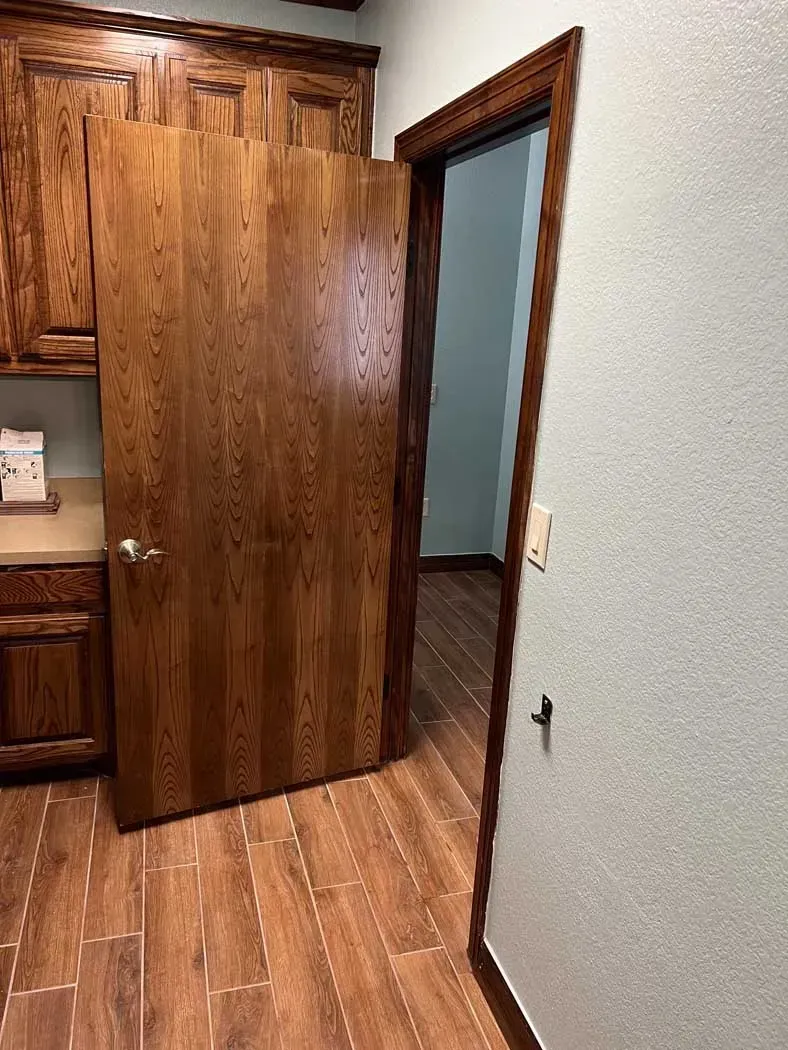 Wooden door ajar in a room with wood-look tile flooring and cabinets. Reveals a light-colored room.