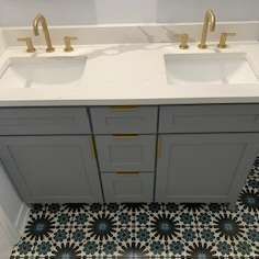 Bathroom vanity with gray cabinets, white countertop, gold faucets, and patterned tile floor.