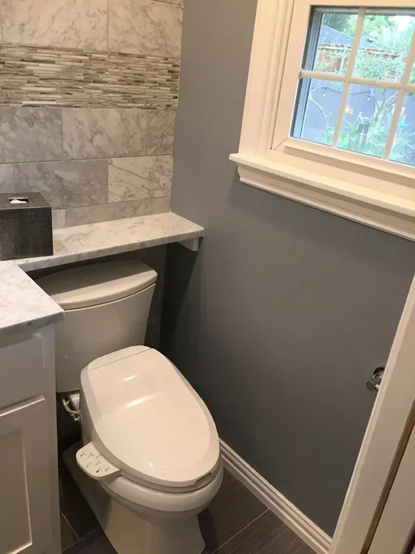Toilet in a gray-painted bathroom with marble tile and shelf, next to a window with white trim.