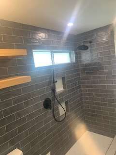 Gray tiled shower with black fixtures, two wooden shelves, and small window.