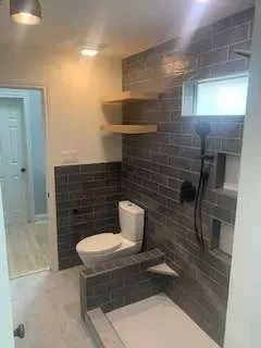 Bathroom with gray tiled shower and toilet, wood shelves, and a white door.