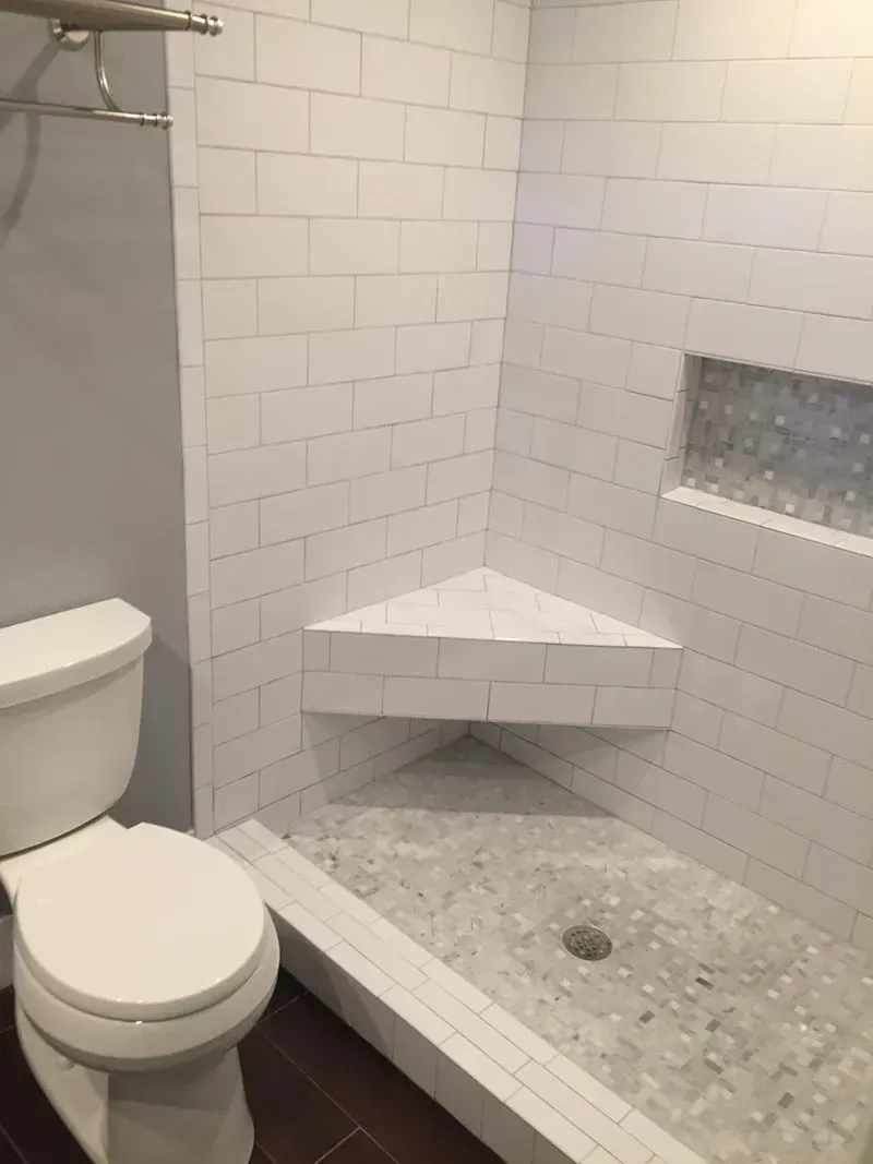White-tiled shower with a corner seat and recessed shelf. Includes toilet and gray wall.