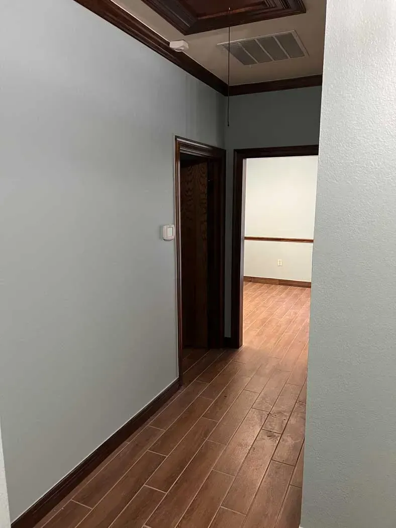Hallway with gray walls, wooden trim, and wood-look flooring, leading to a room.