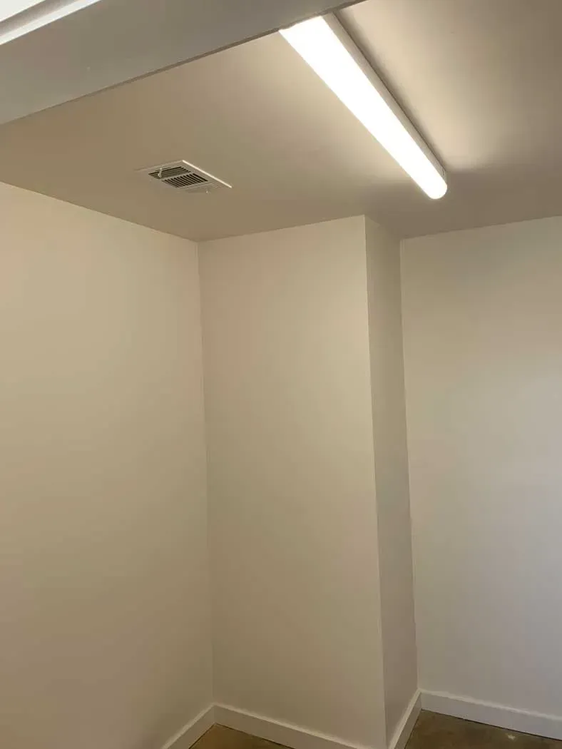 A small, bright room with white walls, a fluorescent light fixture, and a smoke detector on the ceiling.