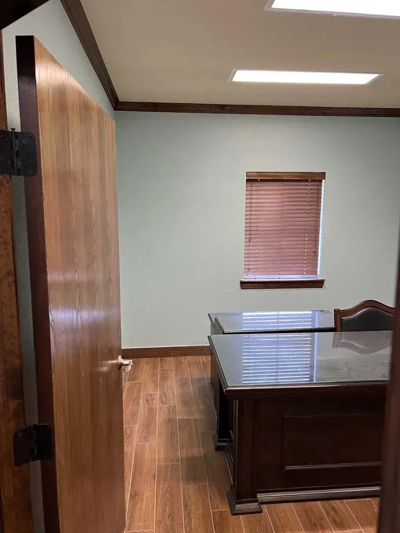 Office interior with wooden door ajar, desk, window with blinds, wooden floors, and blue walls.