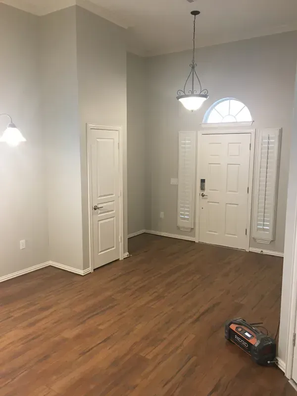 A brightly lit, empty room with wood flooring, white trim, and a high ceiling. A front door is visible.