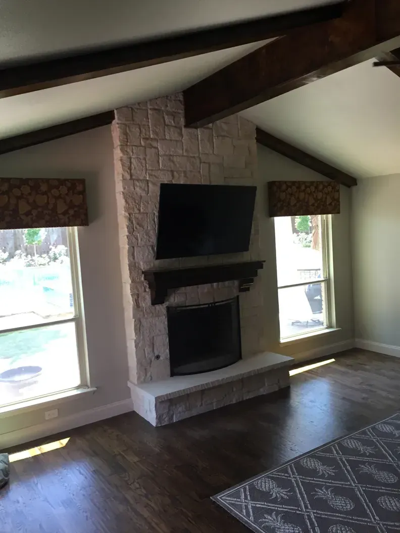 Stone fireplace with mounted TV, windows, wood beams, and patterned rug.