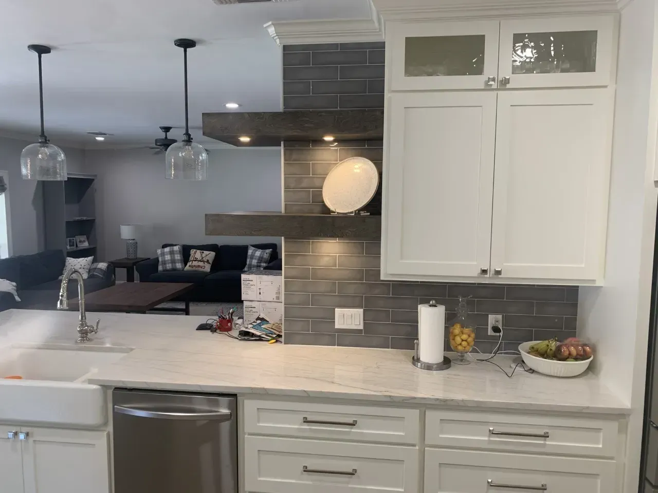 Kitchen with white cabinets, gray backsplash, and open shelving; dining area with black couch in background.