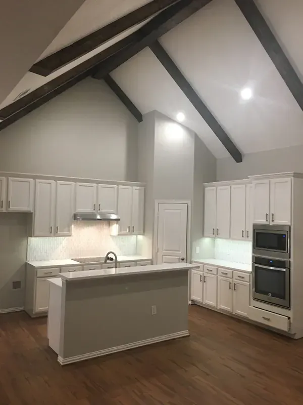 Bright white kitchen with vaulted ceiling and dark wood beams. Island in the center, built-in appliances, and hardwood floors.