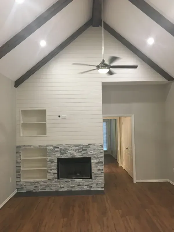 Living room with fireplace, white shiplap wall, grey stone, wooden floors, and a vaulted ceiling with beams.