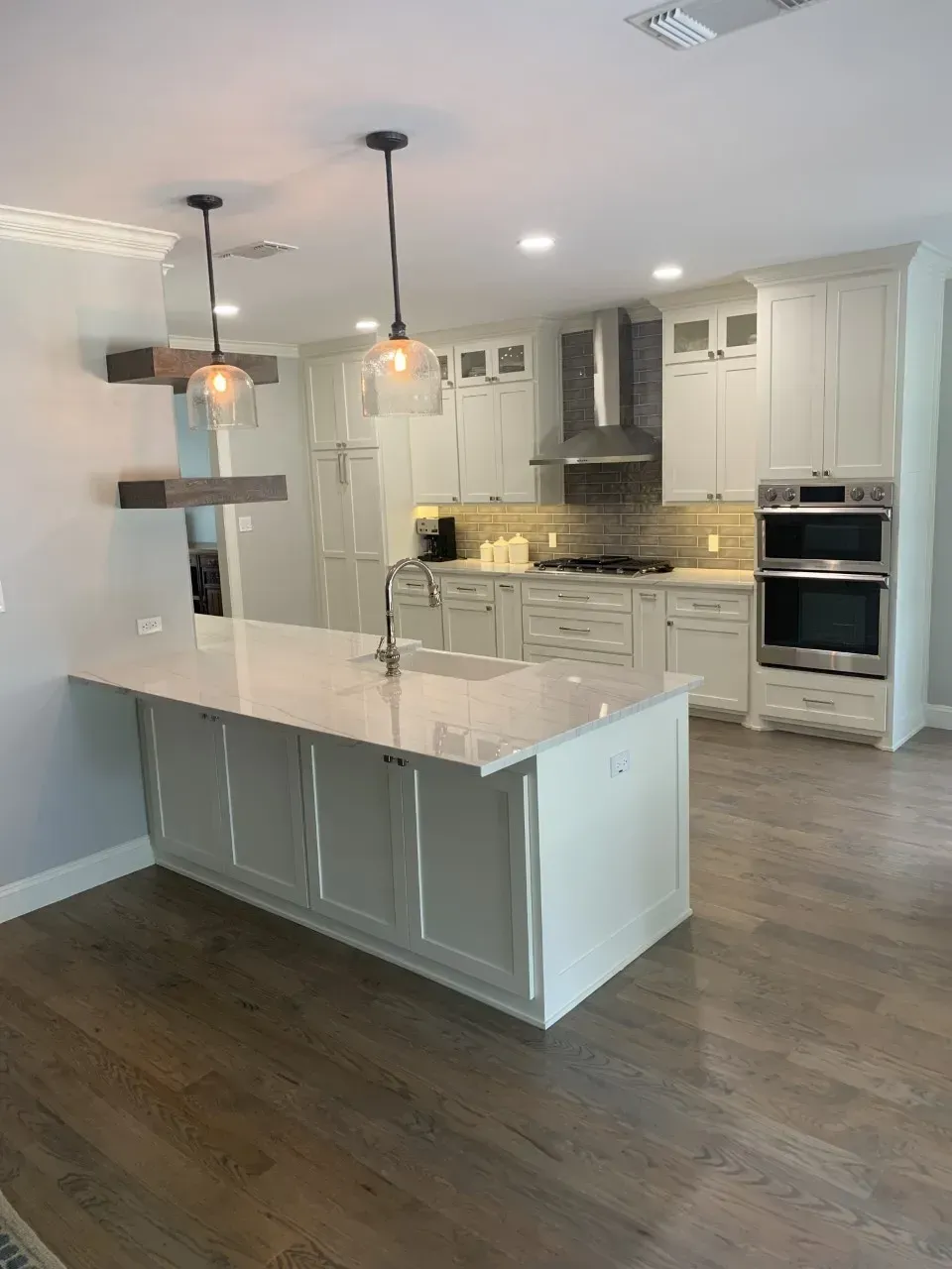 Modern white kitchen with island, pendant lights, stainless steel appliances, and wood floors.