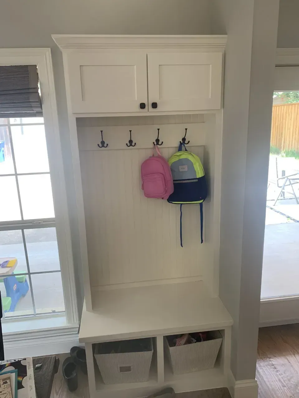 White built-in storage unit with hooks, cabinets, bench, and baskets. Two backpacks hang from hooks.