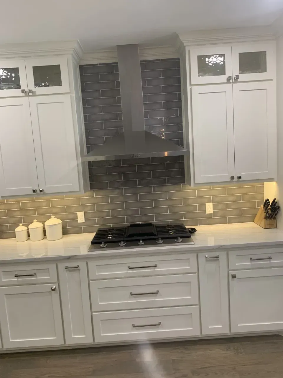 White kitchen with stainless steel range hood, grey tiled backsplash, and white cabinets.