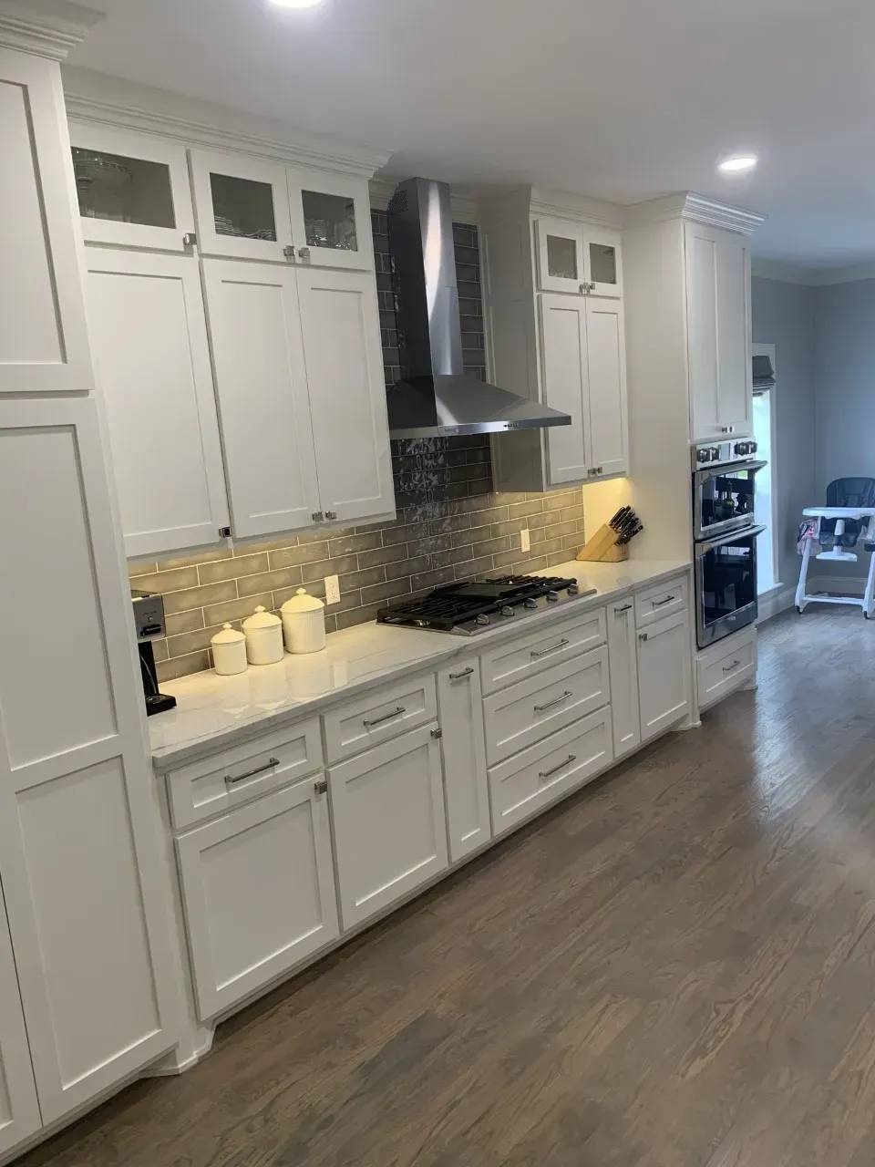 White kitchen with light-colored cabinets, countertops, stainless steel range hood, and tile backsplash.
