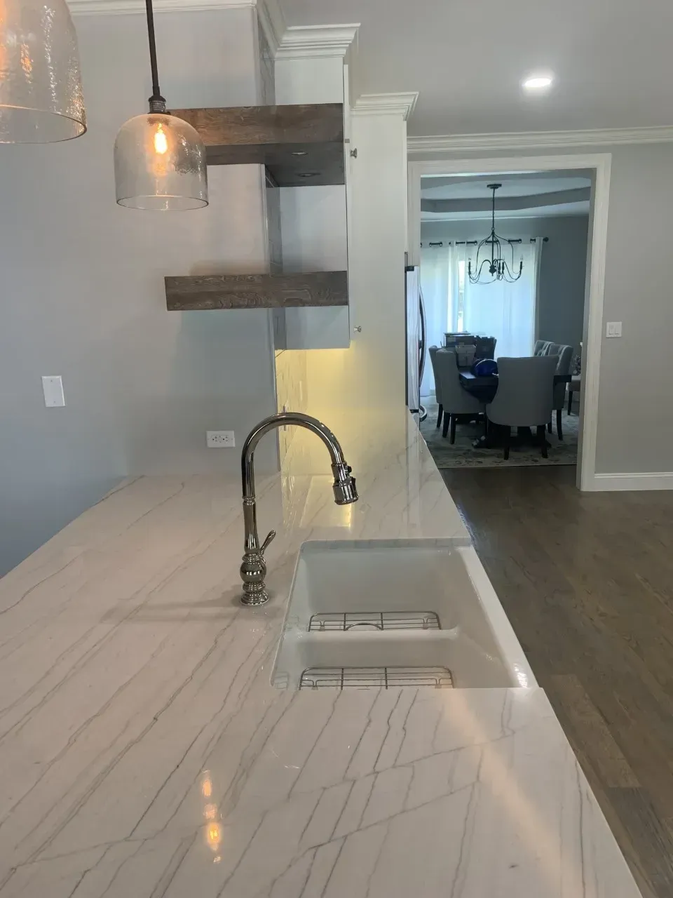 Kitchen countertop with faucet and sink. Open to a dining room. Gray walls and wood shelves.