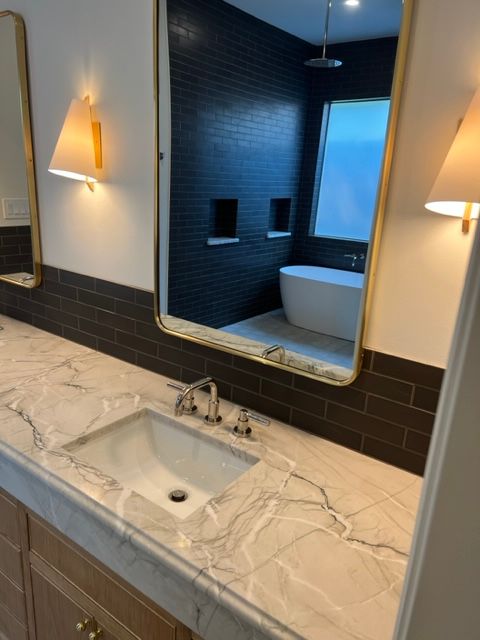 Bathroom with marble countertop, sink, gold-framed mirrors, and dark tiled wall. A tub is visible in the reflection.
