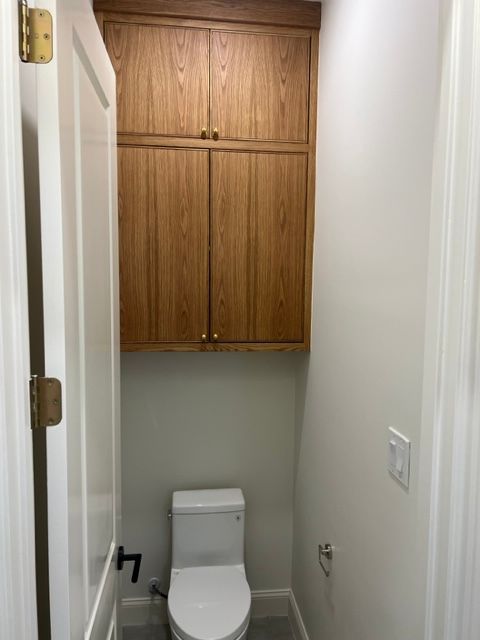 Small bathroom with a toilet and wooden cabinets above. White walls and door.