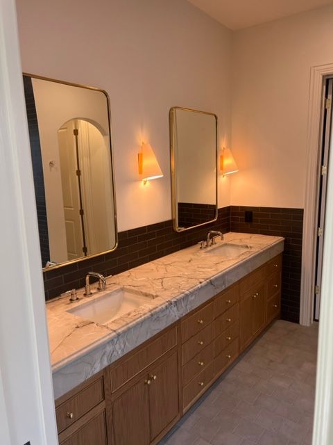 Bathroom with two sinks, two mirrors, and wood cabinets. Dark tile backsplash, light countertops, and gold accents.