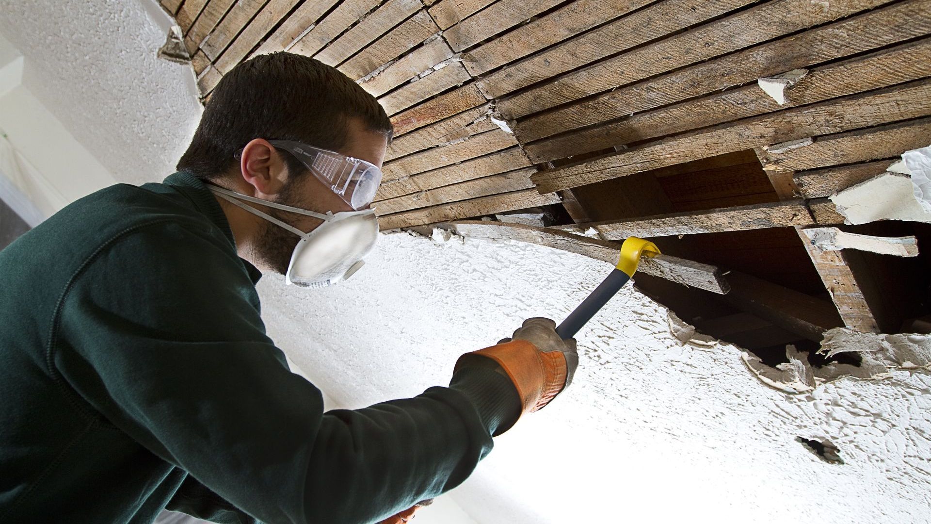 Man in safety gear demolishes ceiling with hammer.
