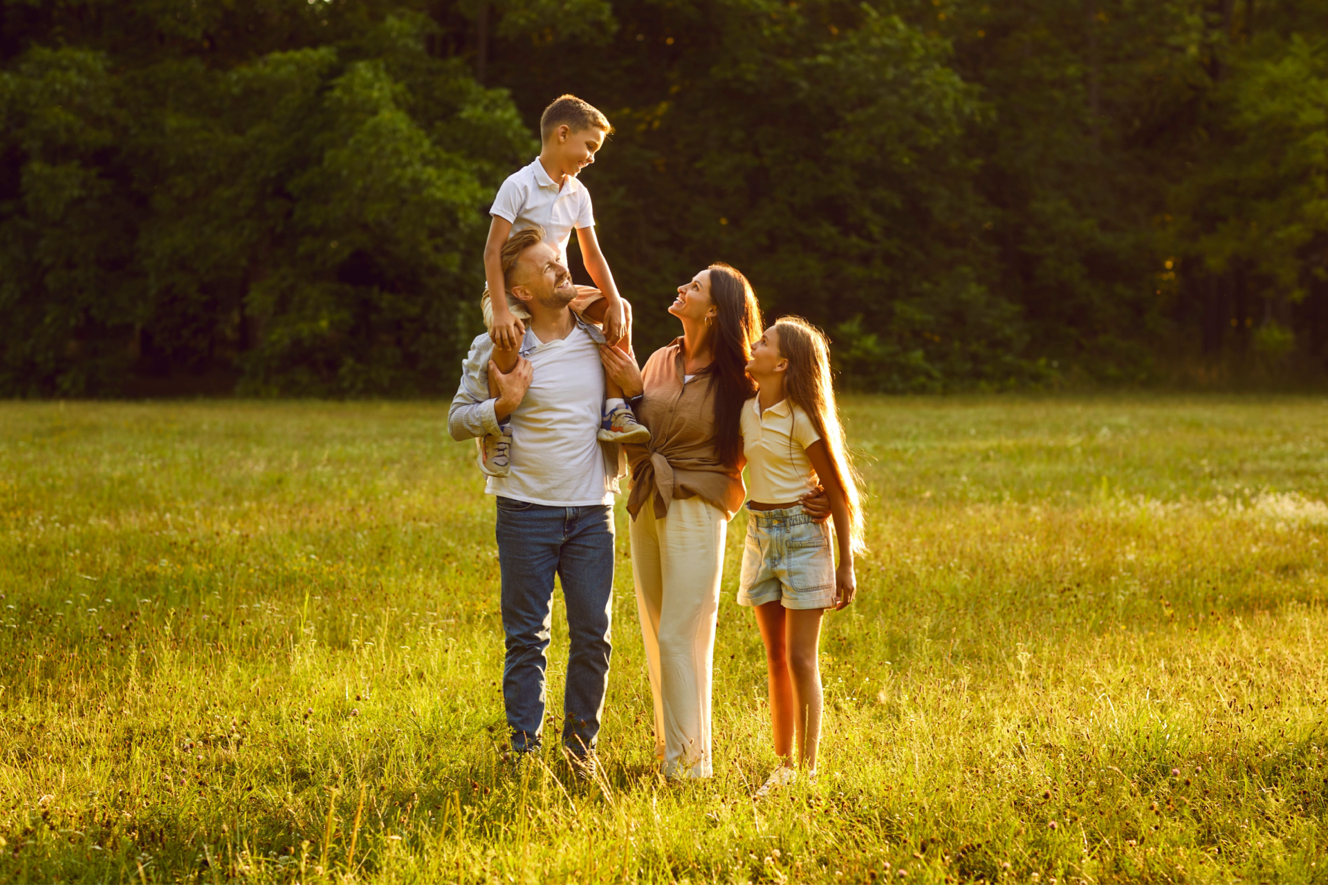 A family walks together in a sunlit field, with a parent carrying a child on their shoulders.