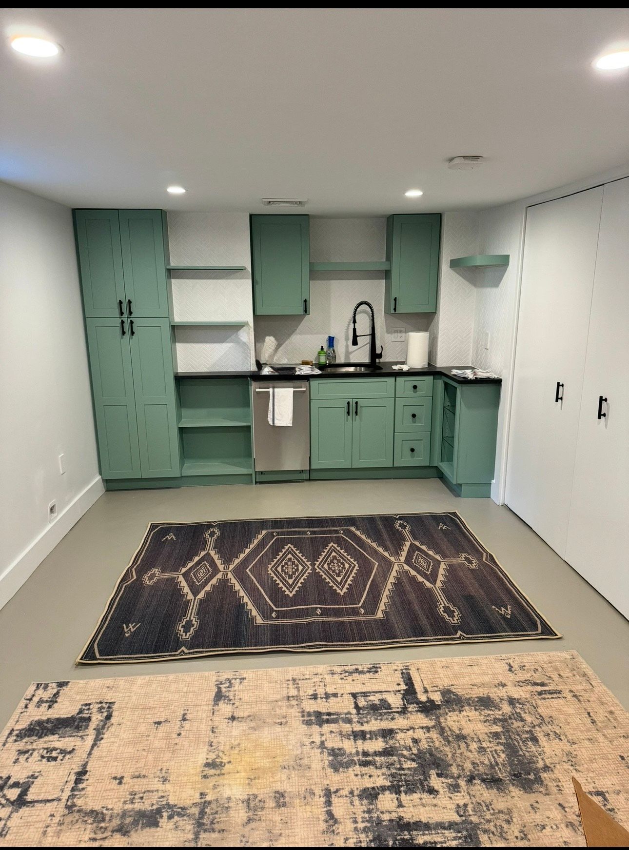 Small kitchen with sage green cabinets, black countertops, and patterned rugs on gray floor.