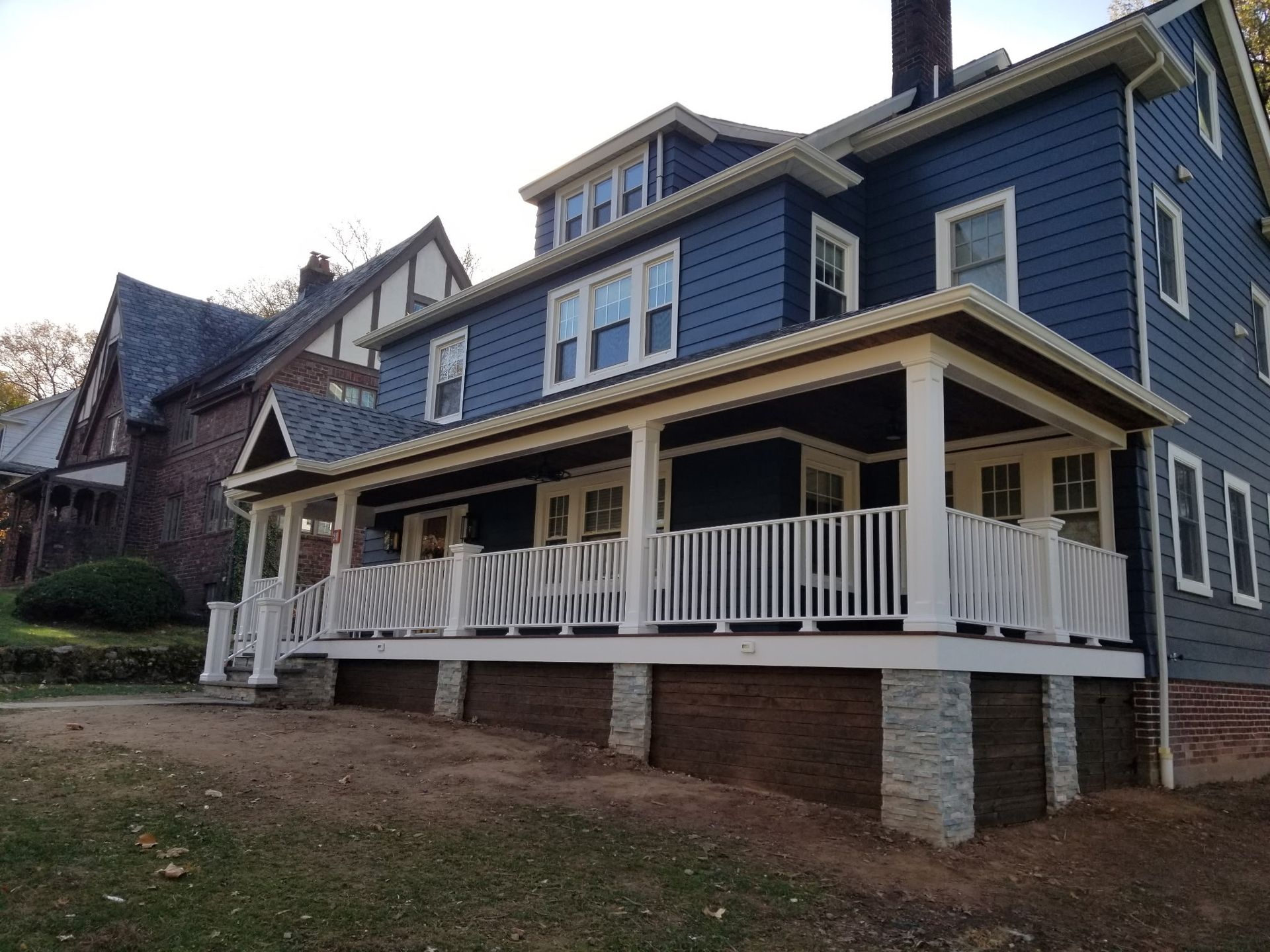 Two-story blue house with a white porch, brick and stone foundation, beside a brick house under a tree.