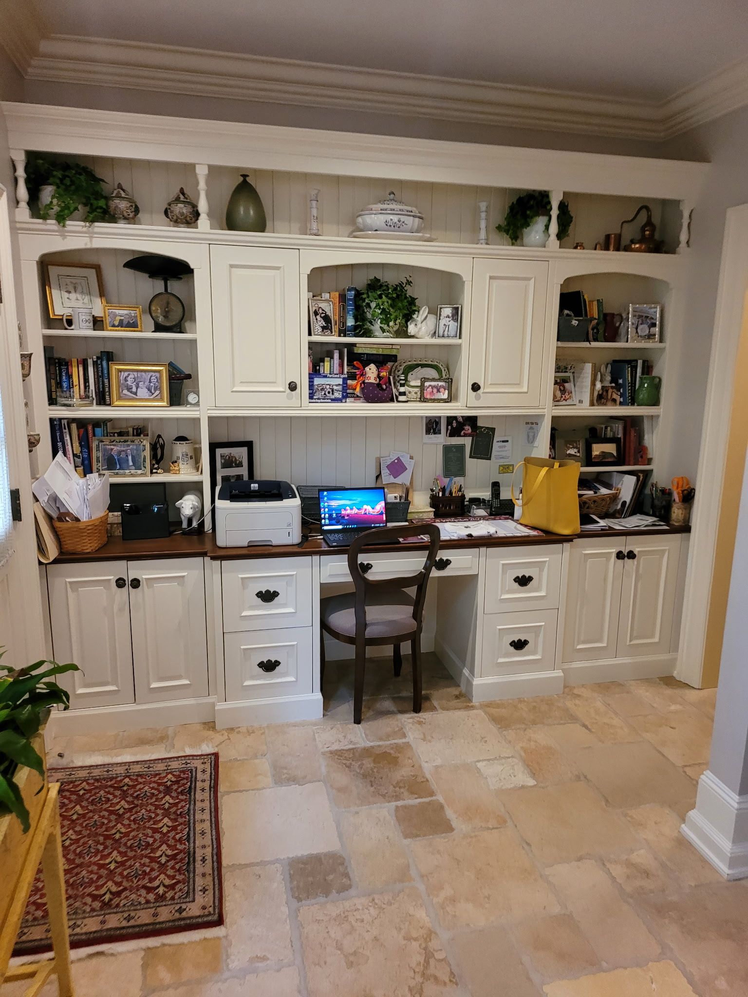 Home office with built-in cream cabinets, desk, chair, and decorations. Stone floor, plants, and rug visible.