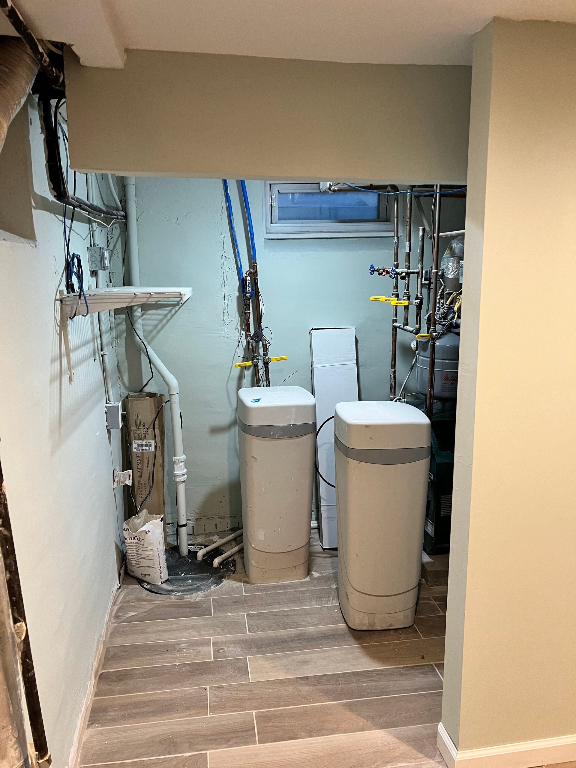 Utility room with water softeners; light green walls, gray flooring, pipes and wiring.