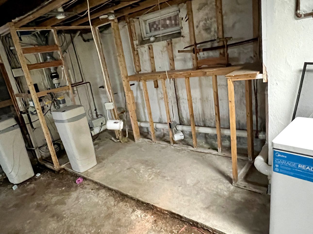 Basement with exposed framing, concrete floor, and pipes. Water softener and a white appliance visible.
