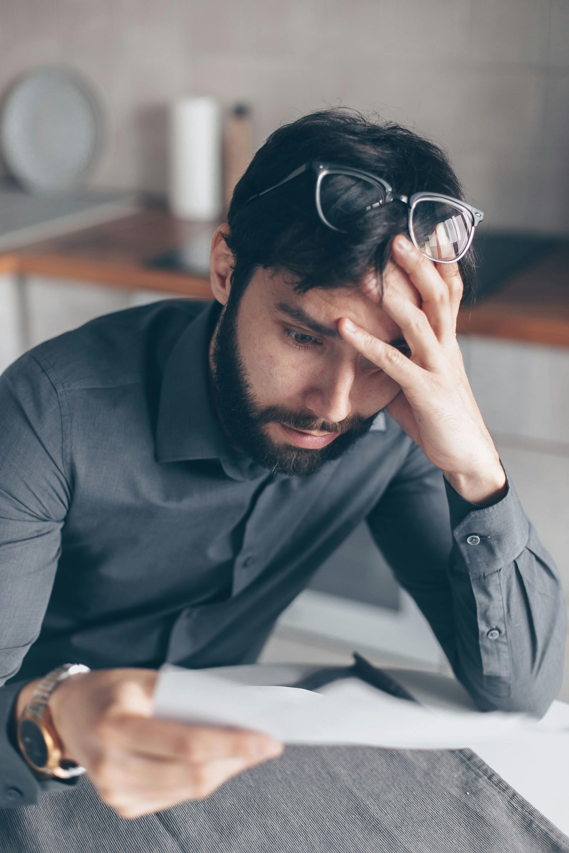Man with hand on forehead, looking down at papers in a kitchen. He appears stressed.