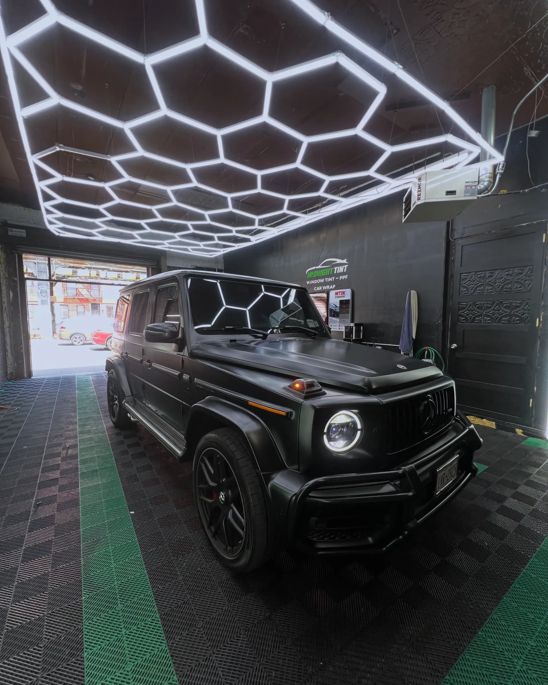 Black Mercedes-Benz G-Wagon parked under hexagon lights inside a car shop with green carpet and open door.
