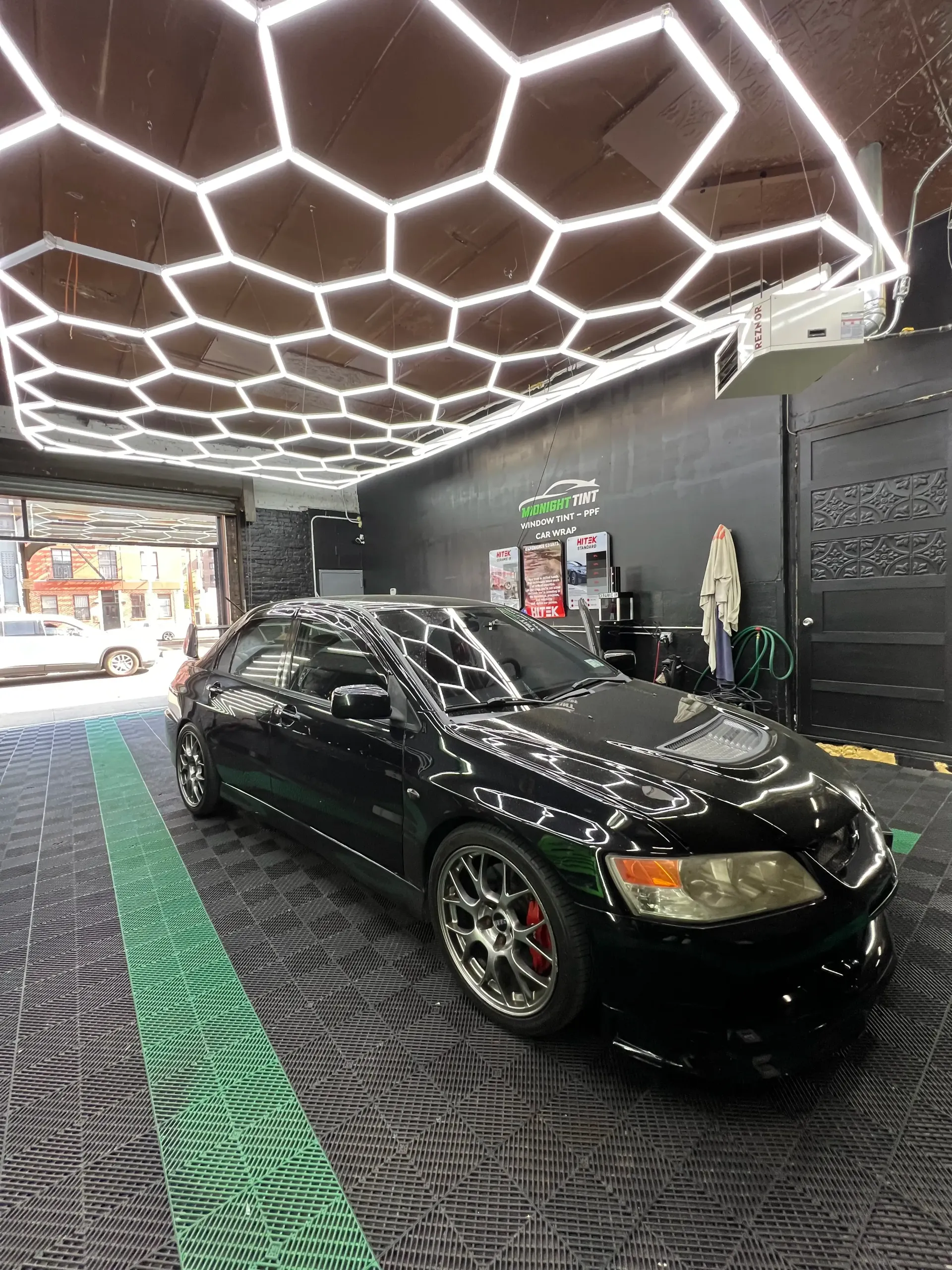 Black car in a detail shop with hexagon-shaped lights overhead, parked on patterned flooring.