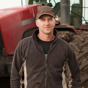 Man in cap and zip-up jacket smiles in front of a red tractor.