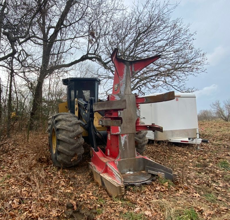 Yellow and black forestry machine with red grapple near a white trailer and trees.