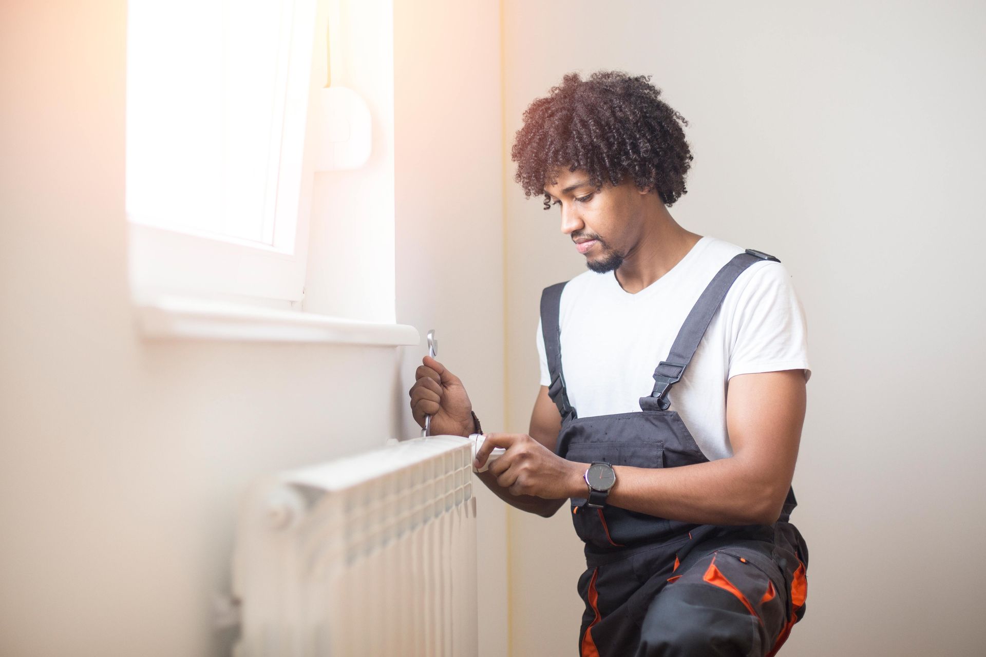 A man in overalls is working on a radiator near a window. He has curly hair and is holding a tool.