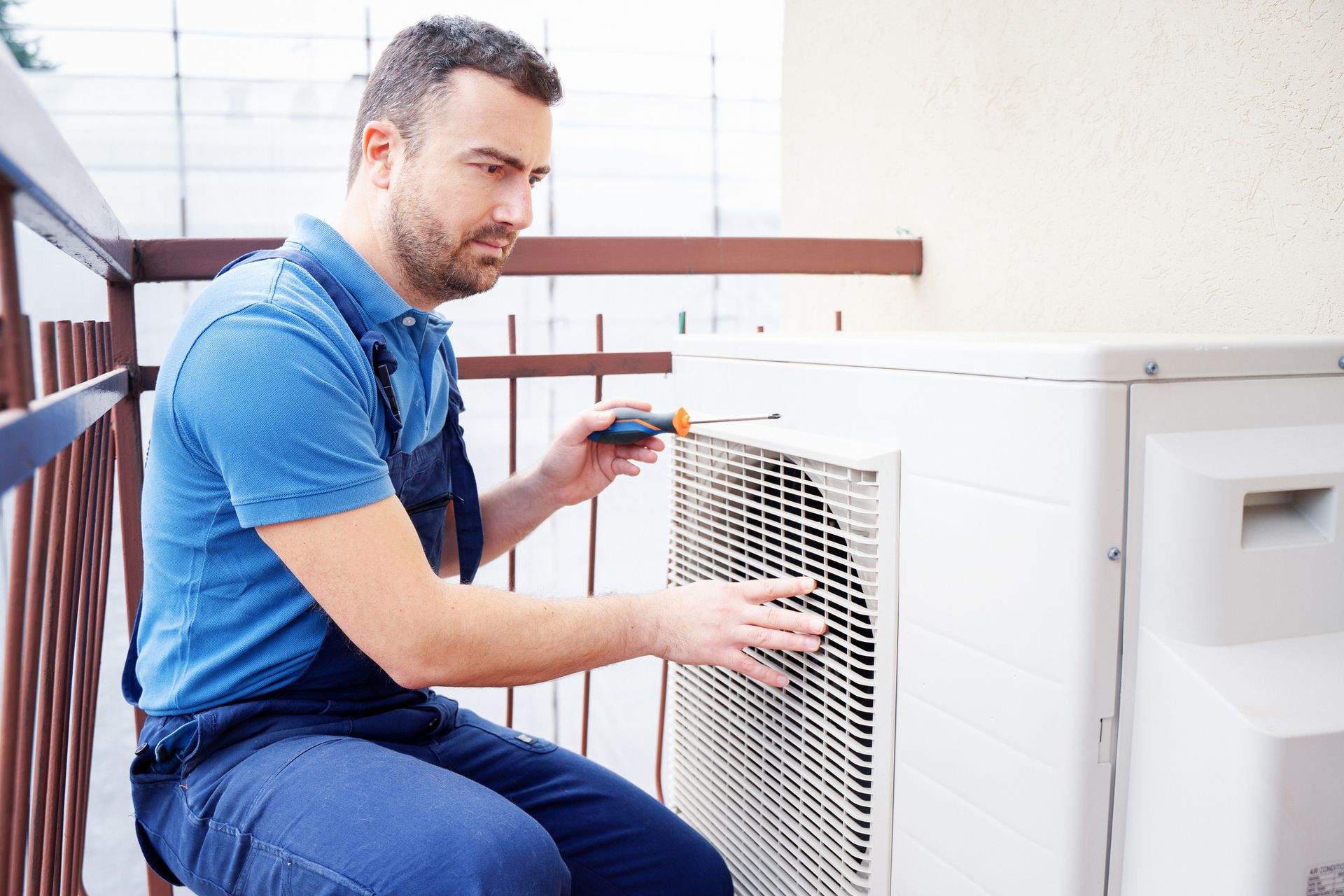 A repairman in blue overalls works on an outdoor AC unit with a screwdriver. He's sitting on a balcony, looking focused.