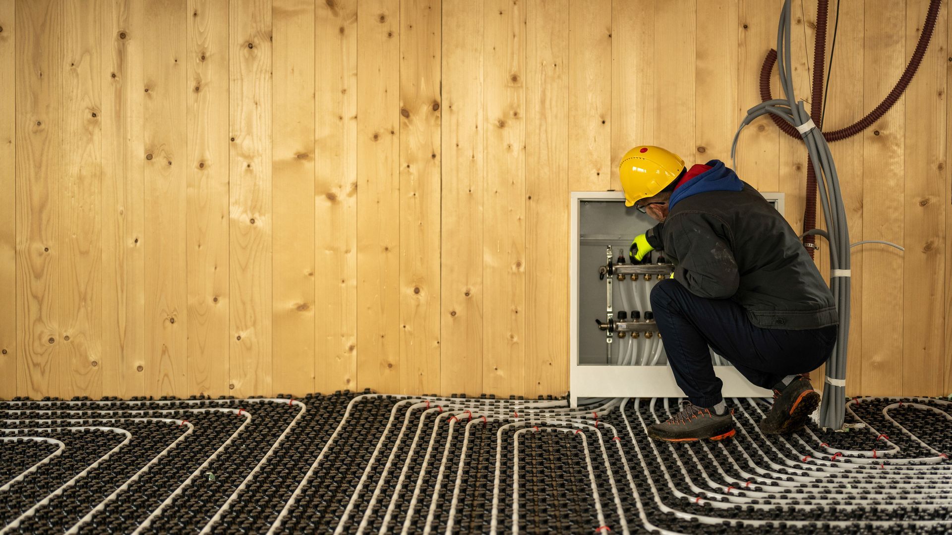 A person in a yellow hard hat inspects pipes in a cabinet, floor heating visible, wood-paneled wall.