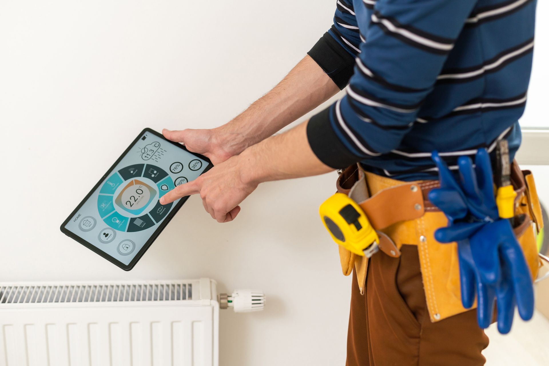 Person adjusts a smart thermostat on a tablet near a radiator. He wears work clothes, including a tool belt and gloves, in a home setting.