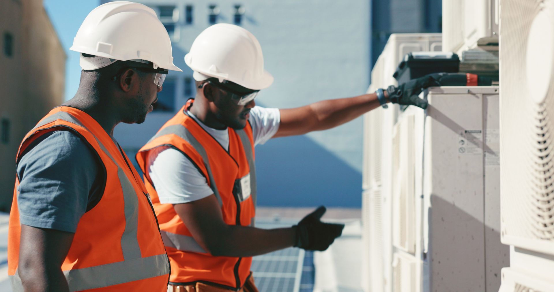 Two construction workers in safety vests and helmets inspect rooftop equipment.