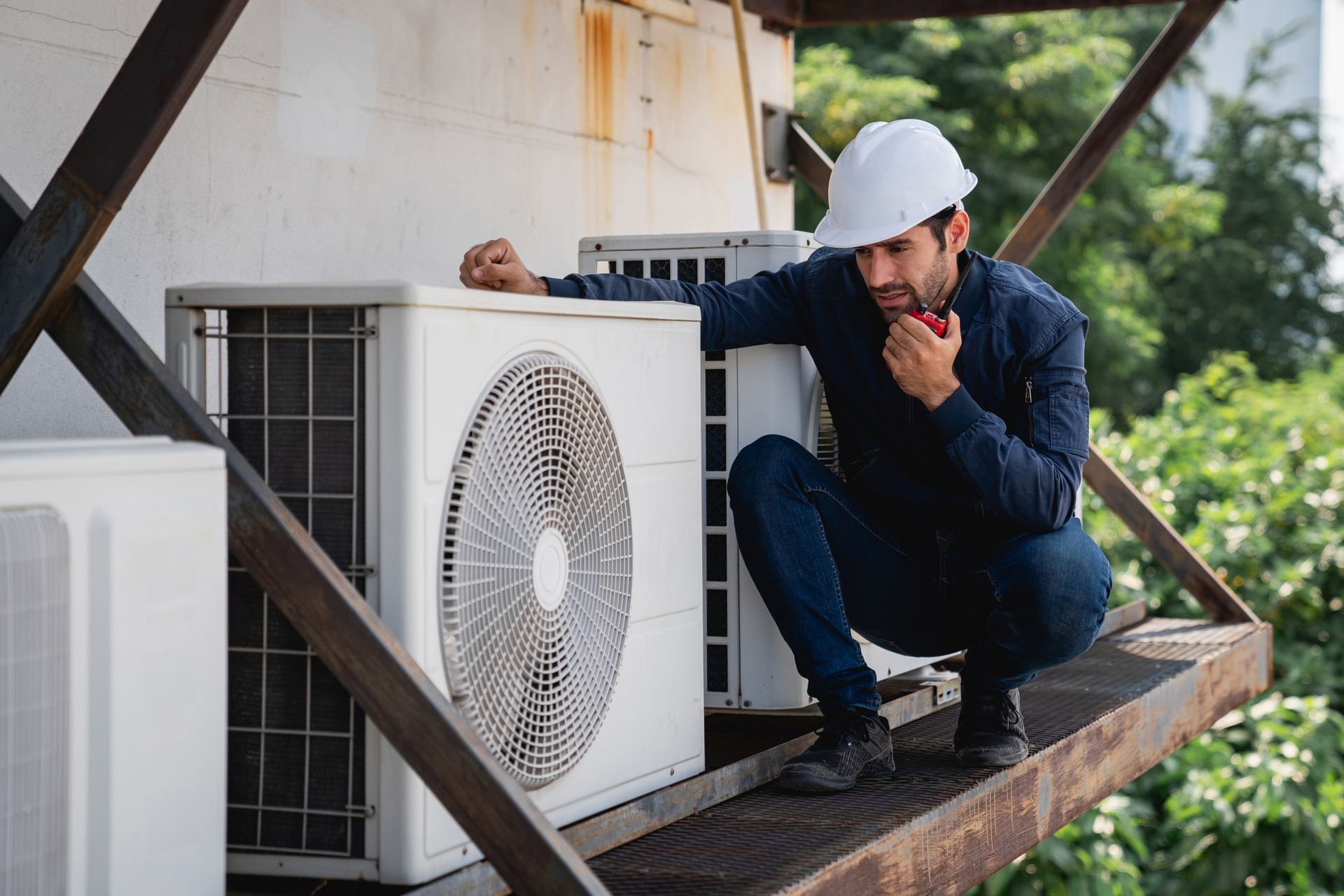 HVAC technician in hard hat inspecting an air conditioning unit on a rooftop. The technician is using a radio and squatting.