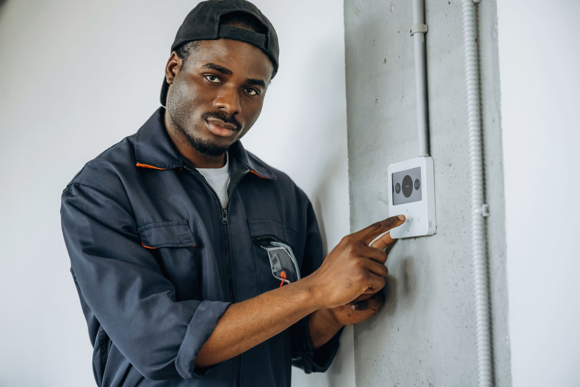 A Black man in a work uniform adjusts a thermostat on a gray wall. He wears a black cap, and looks directly at the viewer.
