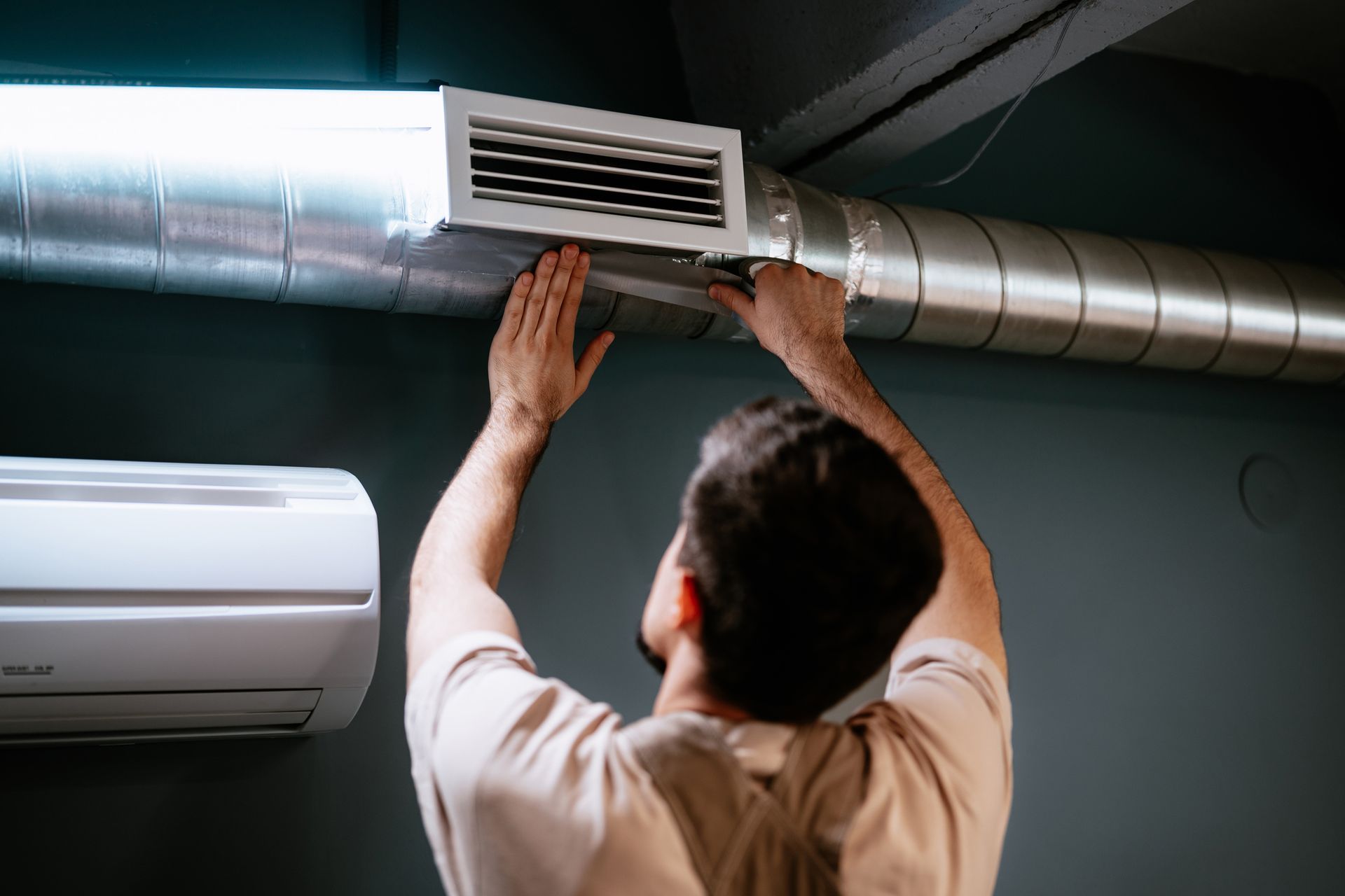 Man installing or adjusting a ventilation vent on a metal duct. An air conditioner is mounted on the wall.