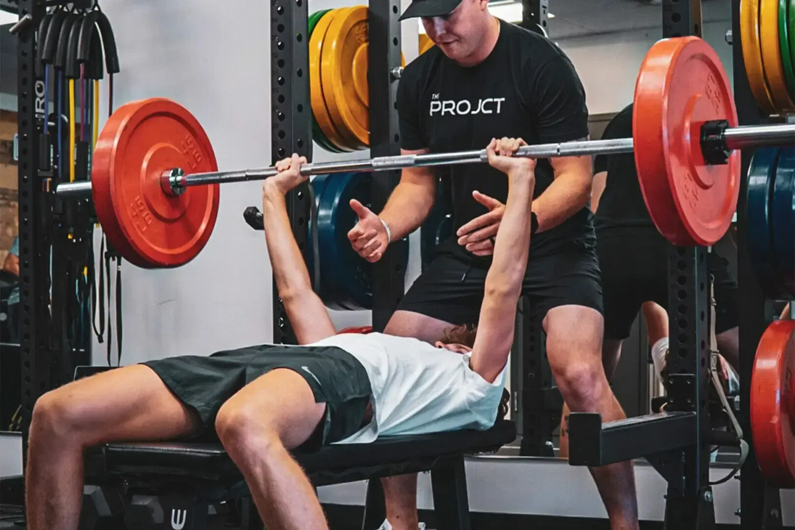 A man is lifting a barbell on a bench in a gym while another man watches.