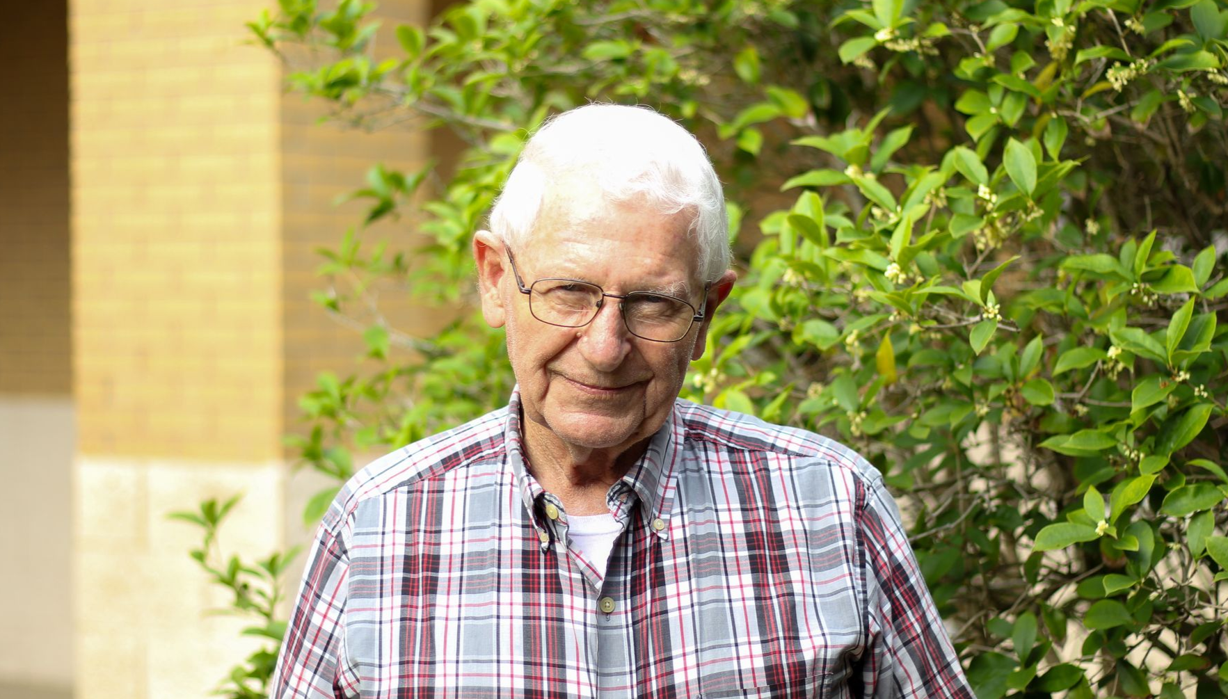 An elderly man wearing glasses and a plaid shirt is standing in front of a church.