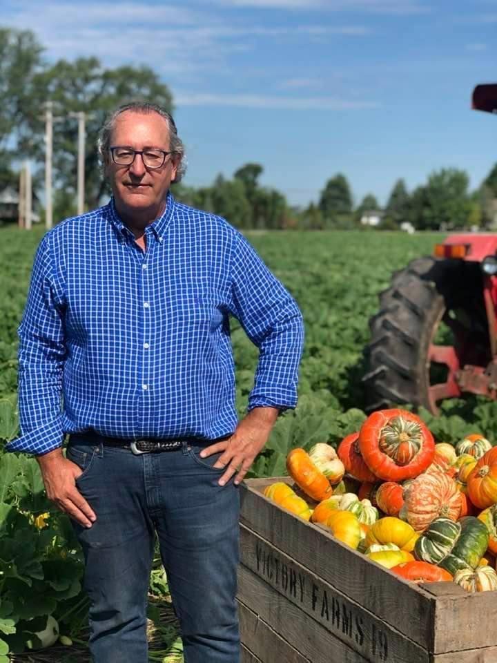Man in blue shirt and jeans stands by a crate of colorful gourds, with a tractor and field in the background.