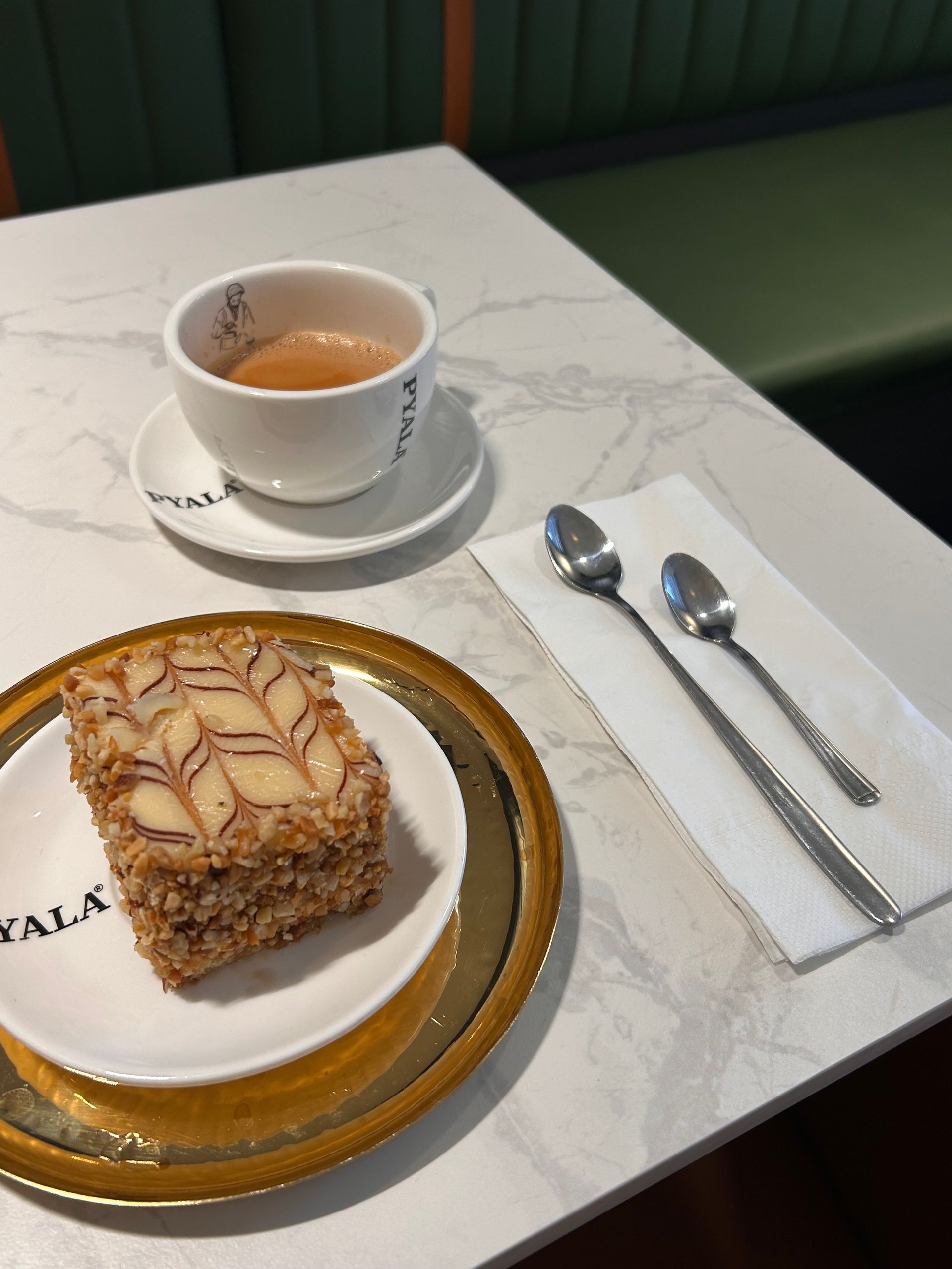 Cup of tea, slice of cake, spoons, and a napkin on a marble table.