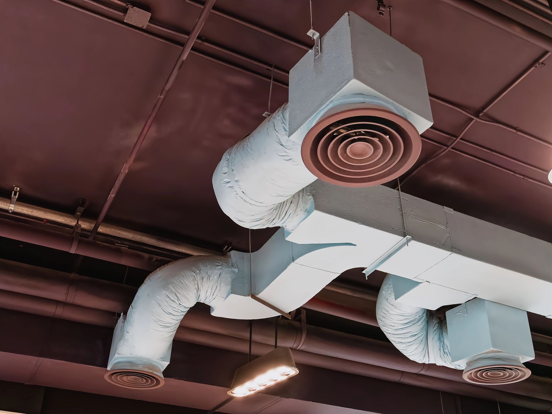Condotti di ventilazione metallici industriali con tre diffusori circolari sospesi da un soffitto marrone scuro.