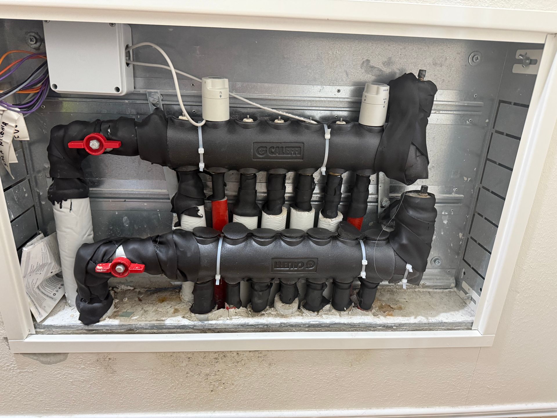 Sistema di distribuzione idronica per riscaldamento con tubi neri isolati, valvole rosse e componenti elettrici bianchi in un armadio a muro.