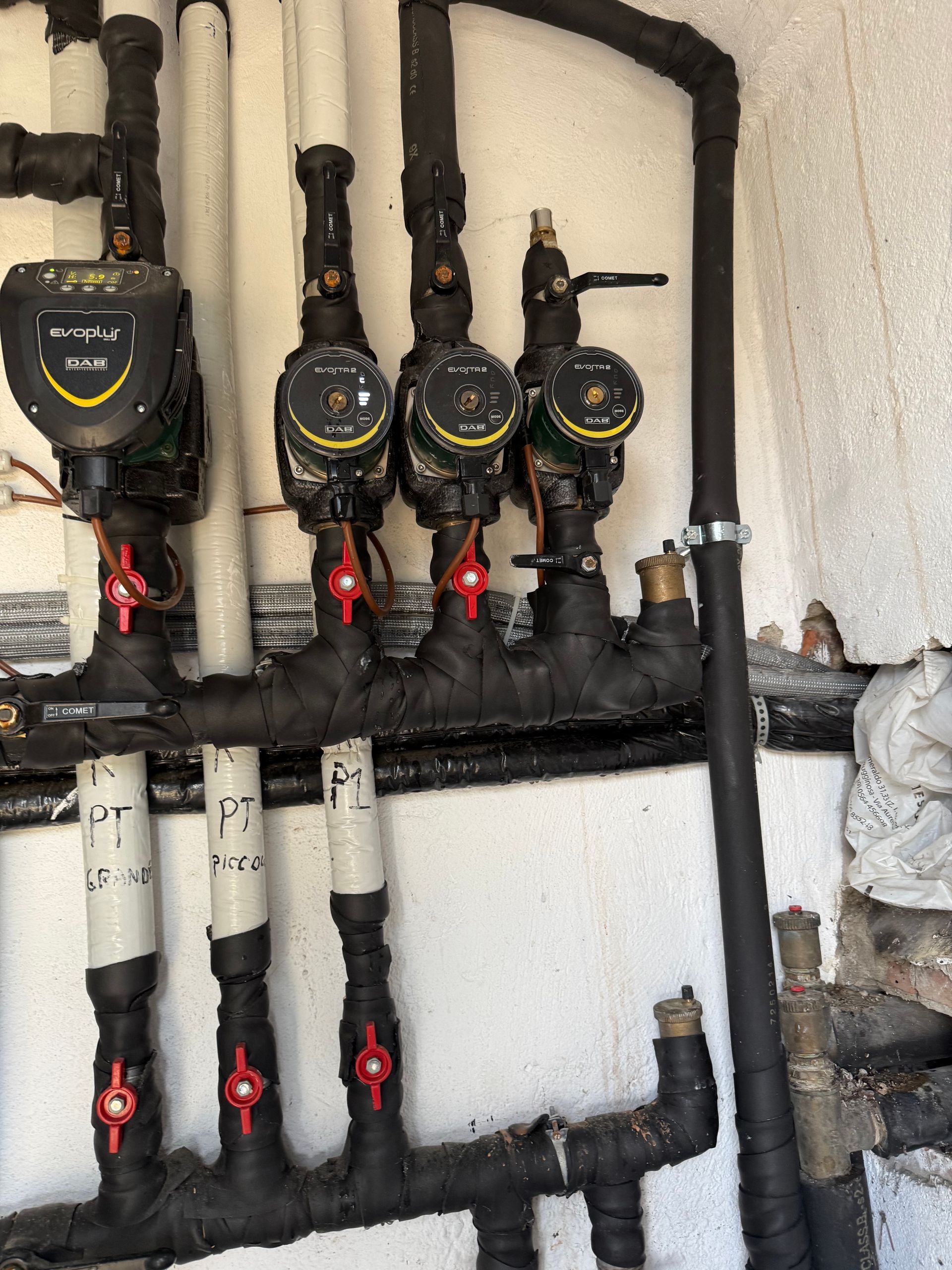 In un locale tecnico, su una parete sono montate quattro pompe di circolazione e tubi con isolamento nero e valvole con maniglia rossa.